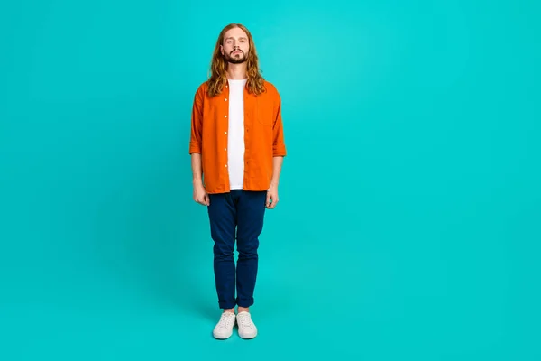 Full length body photo of confident blonde hair guy in orange shirt blue pants standing alone isolated over aquamarine color background.