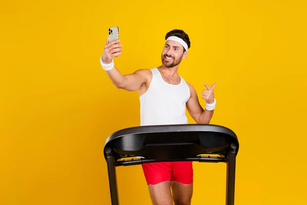 Photo of funky cool guy sportswear running treadmill taking selfie pointing modern device isolated yellow color background.