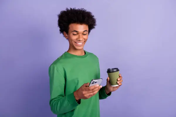 Photo of cheerful funky man dressed green sweater drinking coffee chatting modern device empty space isolated violet color background.