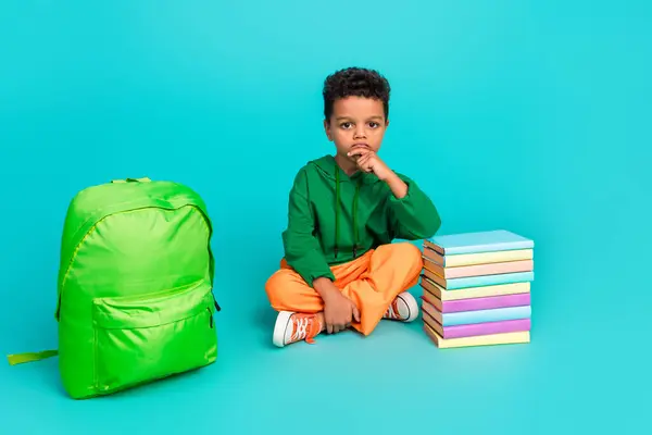 Full size photo of minded boy wear orange pants sit near book backpack finger on chin wait for lesson isolated on teal color background.