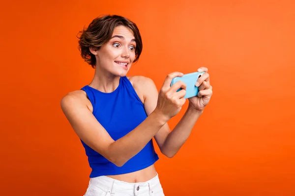 Portrait of funky nervous person biting lips hold smart phone playing games isolated on orange color background.