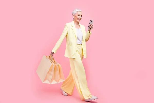 Full body photo of satisfied person dressed jacket walk in empty space hold bags look at smartphone isolated on pink color background.