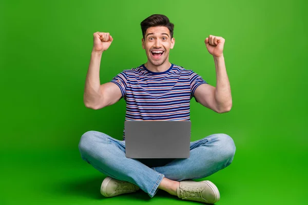 Full length photo of impressed man wear stylish t-shirt sitting on floor with laptop on legs win bet isolated on green color background.