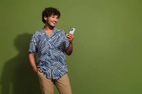 Photo of cheerful positive guy wear print shirt texting modern device empty space isolated khaki color background.