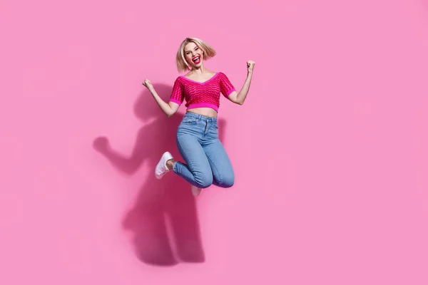 ピンクの背景で孤立したギャンブルを勝ち取るクレンチングフィストジャンプニットトップジーンズパンツを楽しみ狂った女性のフルサイズの写真 — ストック写真