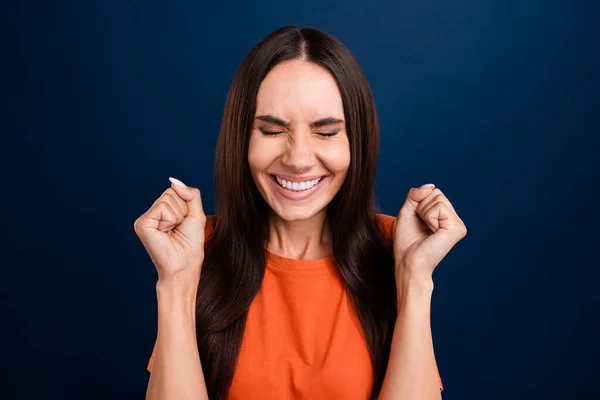 Photo of lucky excited lady wear orange t-shirt closed eyes rising fists isolated dark blue color background.