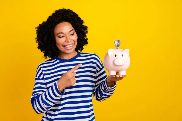 Portrait of cute optimistic girl with chevelure wear striped pullover look directing at money box isolated on yellow color background.