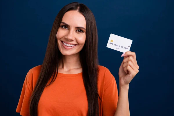 Photo of smart positive woman with straight hairdo dressed orange t-shirt presenting debit card in arm isolated on dark blue background.