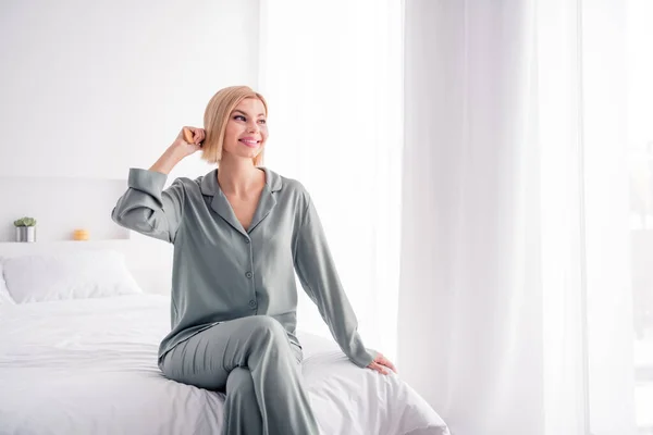 Photo of young dreaming positive satisfied cute girl in pajama sitting on bed indoors light interior bedroom brushing her blonde hair.