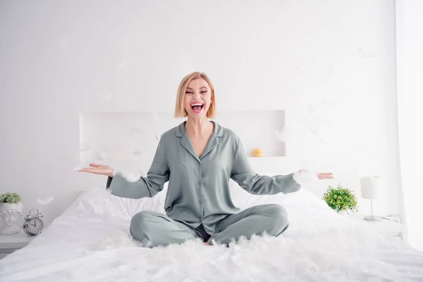 Photo portrait of lovely blonde young lady sit bed excited flying feather wear trendy gray sleepwear comfortable bedroom daytime.