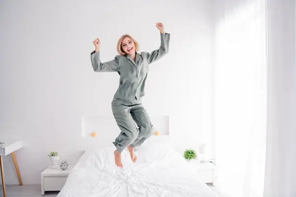 Photo portrait of pretty blonde young girl jump bed have fun celebrate wear trendy gray nightwear bright bedroom interior design.