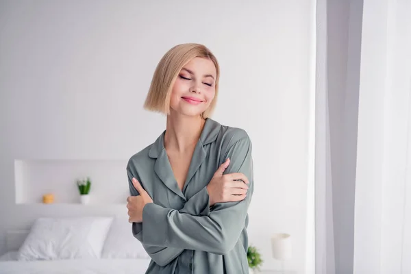 Photo of young beautiful blonde bob hair lady wearing soft grey nightwear sleepy mood hugging herself isolated at home light white interior.