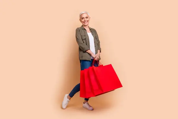 魅力的な引退した女性のフルボディ写真は ベージュ色の背景に隔離されたスタイリッシュなカーキの服を着たショッピングバッグを保持します — ストック写真