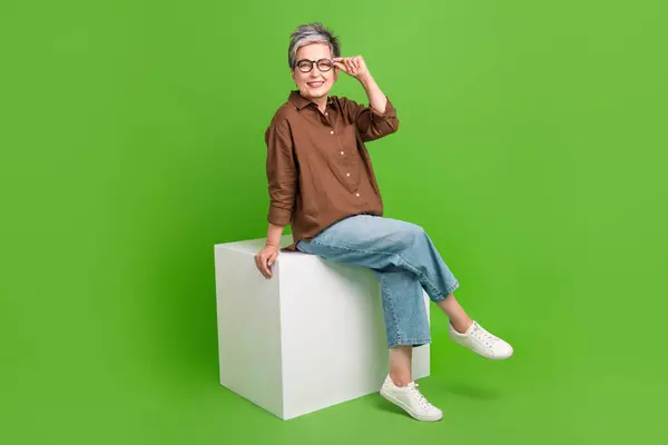 グリーンカラーの背景に隔離されたキューブタッチグラスに座るオーバーサイズのシャツを着た賢い年金女性の全長の写真 — ストック写真