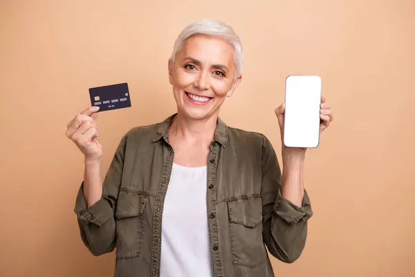 Photo of digital banking features user mature age smiling woman enjoy eshopping hold card and phone isolated on beige color background.