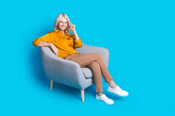ブロンド年金女性のフルボディ写真は 青色の背景に隔離されたスタイリッシュな黄色のサテン服を着た自信を持って椅子に座ります — ストック写真