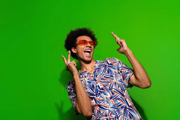Photo of excited funky guy wear print shirt dark eyeglasses dancing showing v-signs empty space isolated green color background.