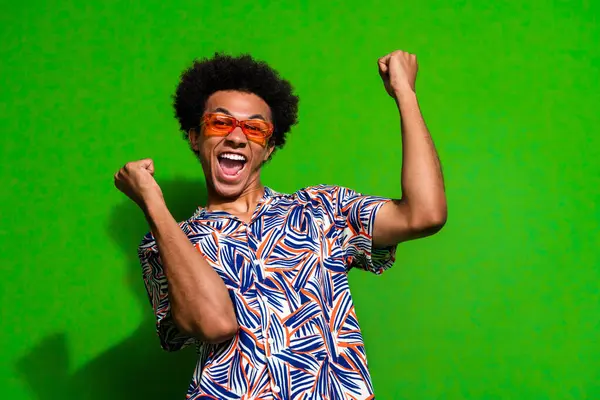 Photo of cheerful positive guy wear print shirt dark eyeglasses yelling yes rising fists empty space isolated green color background.