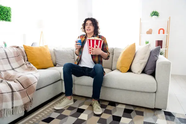 Full length photo of excited funny guy wear plaid shirt eating pop corn can coke watching interesting movie indoors room home house.