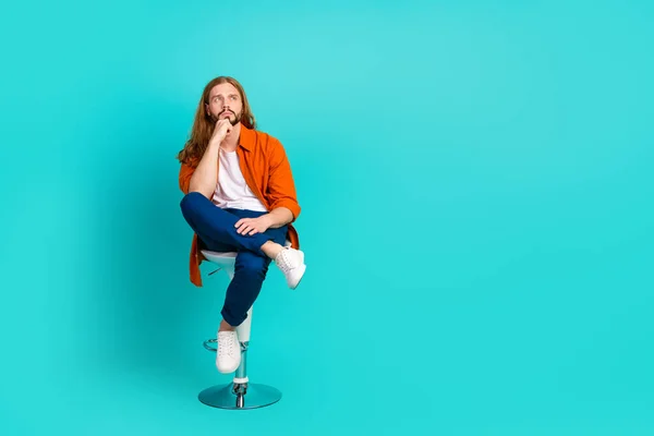 Full length photo of clever man dressed orange shirt sit on chair hand on chin look at offer empty space isolated on teal color background.