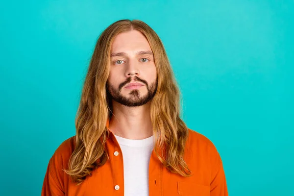 Portrait of young guy with long straight blonde hair wearing orange shirt seems really serious isolated on aquamarine color background.