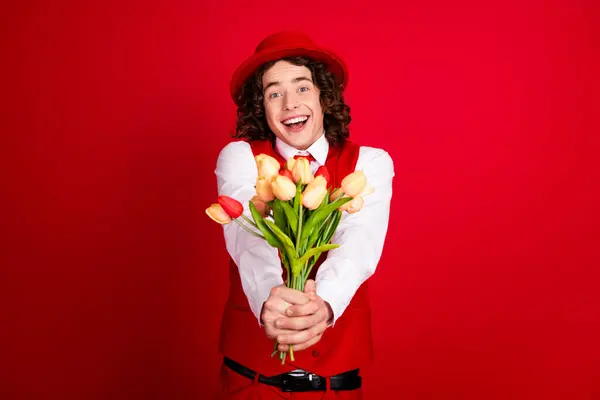 Photo portrait of cute positive guy wearing vintage gentleman outfit greeting you to give tulips bouquet isolated on red color background.