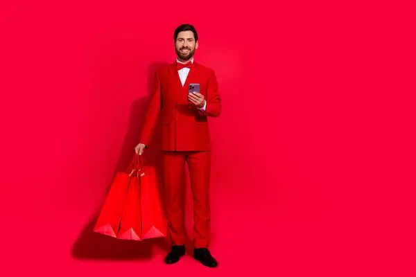 Full body portrait of positive classy guy hold shop bags use smart phone eshop empty space isolated on red color background.