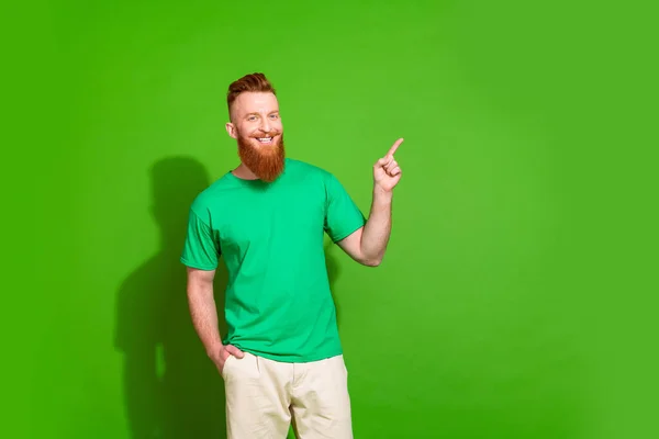 Photo of optimistic man with ginger beard dressed green t-shirt indicating at offer empty space isolated on green color background.