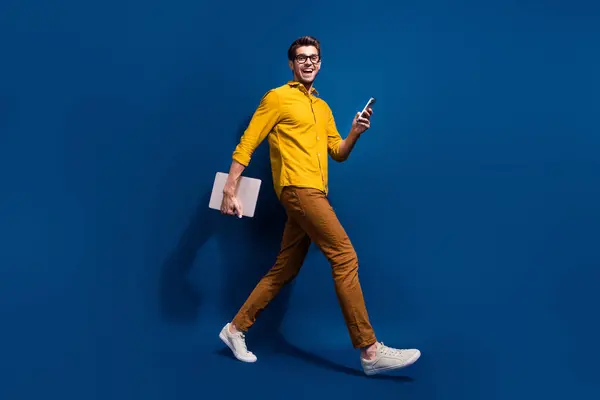 Full body photo of attractive young guy walk netbook device marketer dressed stylish yellow clothes isolated on dark blue color background.