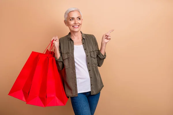 赤いパッケージを保持している短い灰色の髪の女性カーキシャツの写真 ベージュ色の背景に隔離された直接指空のスペースの買い物袋 — ストック写真