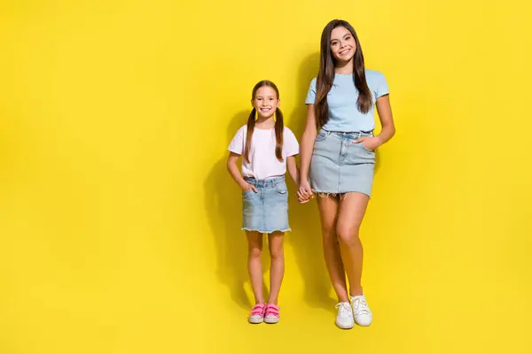 Full length photo of pretty good mood small siblings dressed t-shirts holding hands arms empty space isolated yellow color background.