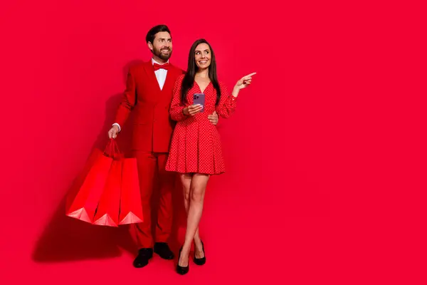两个漂亮情人拥抱购物袋的全长肖像智能手机显示出手指空旷的地方与红色背景隔离 — 图库照片