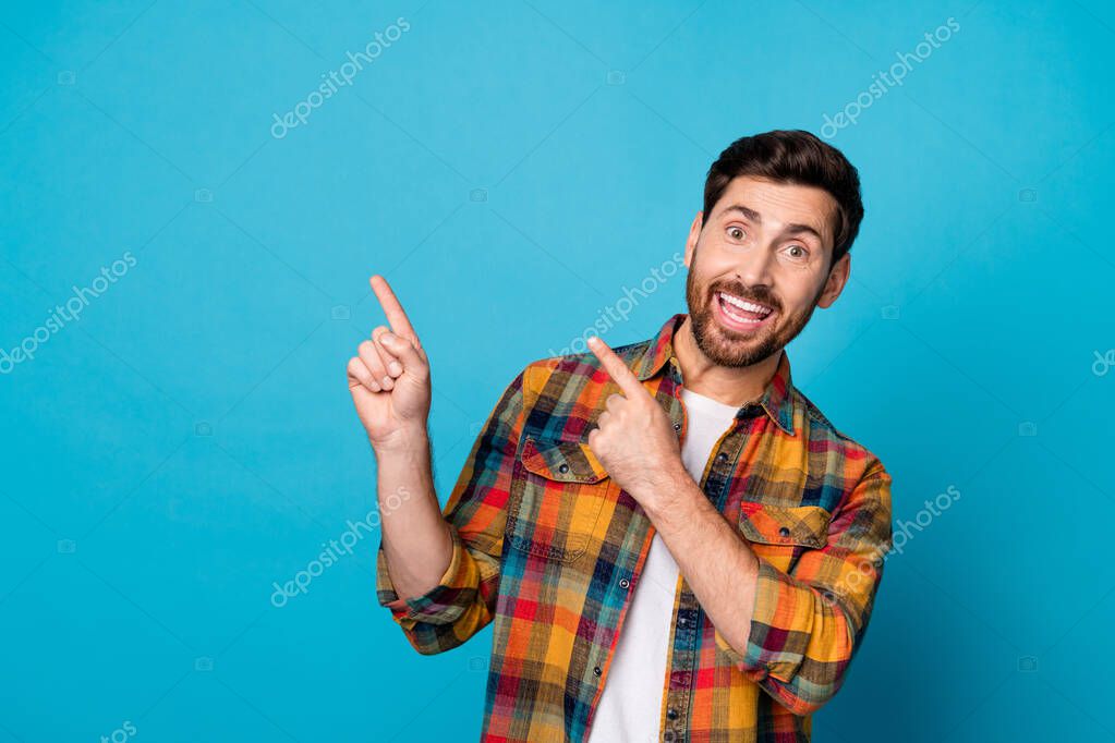 Photo of toothy beaming man with beard dressed checkered shirt indicating at promotion empty space isolated on blue color background.