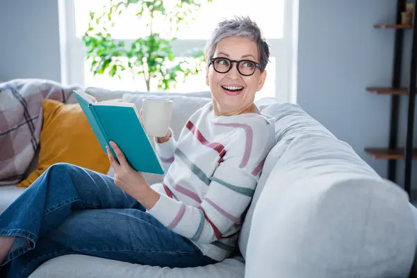Profile portrait of positive aged person sitting sofa hold opened book coffee mug look interested away daylight flat indoors.