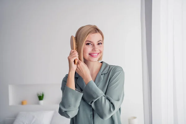 Portrait of sweet cute young lady wear grey nightwear wake up and brushing her blonde hair with comb in light interior house indoors.