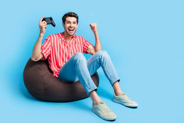満足した男性のフルサイズの写真は ビーンバッグに座ってストライプシャツを着用します ジョイスティックウィンステーションゲーム ブルーカラーの背景で隔離 — ストック写真
