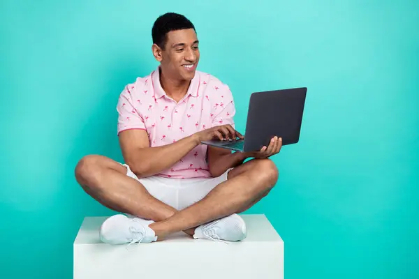 聪明男子身穿粉色马球白色短裤坐在立方体上的全长照片 在绿松石色背景的臂膀下 看着笔记本电脑 — 图库照片