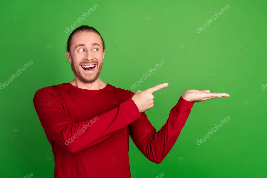 Portrait of ecstatic guy with long hair wear red shirt directing staring at product on palm empty space isolated on green color background.