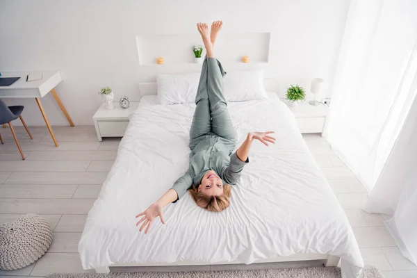 Full body length photo of relaxed blonde girl have rest in bedroom lying down bed legs upside raised hands up when wake up at home.