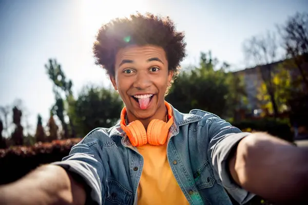 Photo of funky carefree childish youngster dressed jeans jacket with headphones on neck making selfie stick out tongue in park outdoors.
