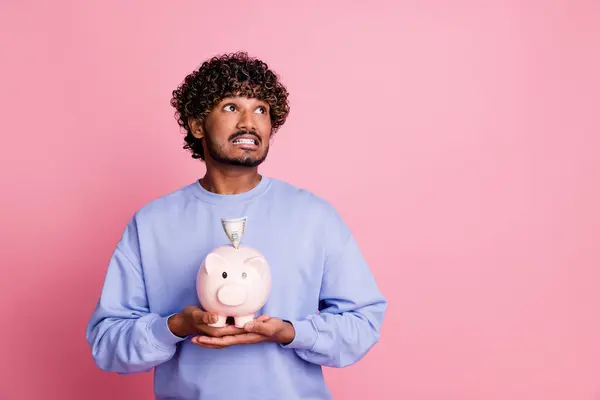 Photo of worried nervous man hold piggy bank collect cash banknotes deposit credit look up empty space isolated on pink color background.