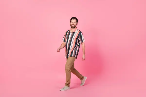 ピンク色の背景に隔離された夏の服を散策するストライプシャツと茶色のズボンの陽気な男の全身の長さの写真 — ストック写真