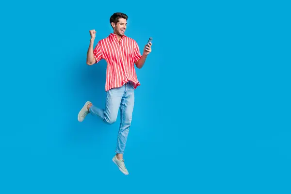 全尺寸照片满意的男人穿条纹衬衫斜纹棉裤跳跃看智能手机赢赌蓝色背景孤立 — 图库照片
