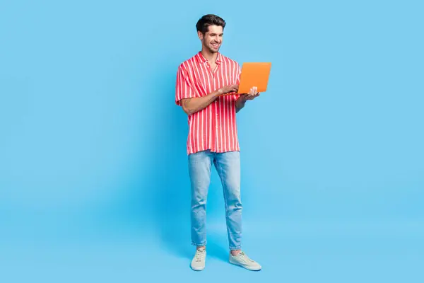 聪明而专注的男人穿着时髦的T恤牛仔裤长裤的全身像看笔记本电脑 用蓝色背景把文章隔开 — 图库照片