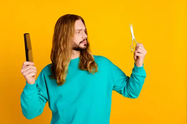 Photo of minded person look curious hand hold hair comb scissors decide ponder isolated on yellow color background.
