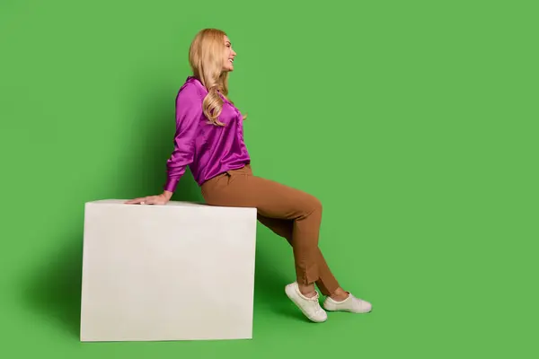 夢のような光沢のある女性の完全な長さの写真は 白い表彰台に座ってヴァイオレットシャツを着用し 空白のスペースを隔離された緑色の背景を見ます — ストック写真