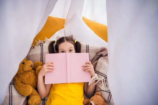 可爱小女孩躺在地板上的照片肖像帐篷盖着脸看书穿上时髦的黄色衣服现代游戏室的内部 — 图库照片