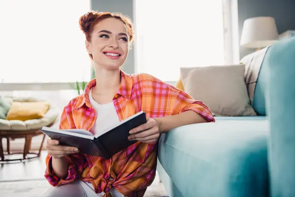 Portrait of gorgeous positive minded girl hold opened book toothy smile fantasize enjoy free time apartment inside.
