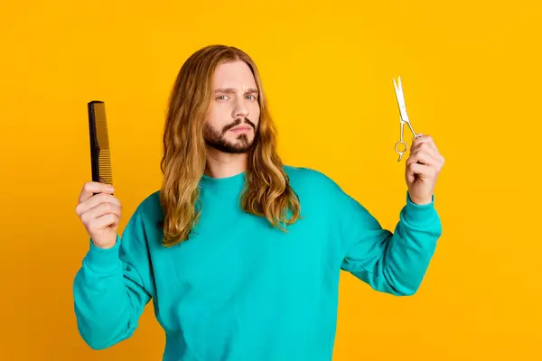 Portrait of questioned minded man arms hold comb scissors contemplate isolated on yellow color background.
