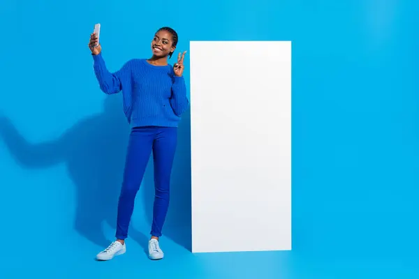 Photo of pretty funky woman wear sweatshirt tacking selfie device v-sign empty space isolated blue color background.